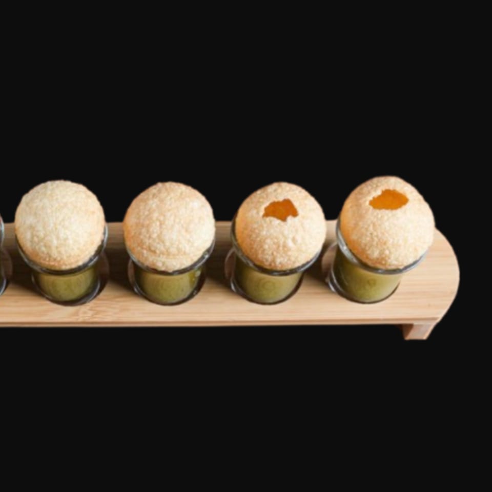 Four pani puri shots with green liquid, two with filling visible, on a wooden tray.